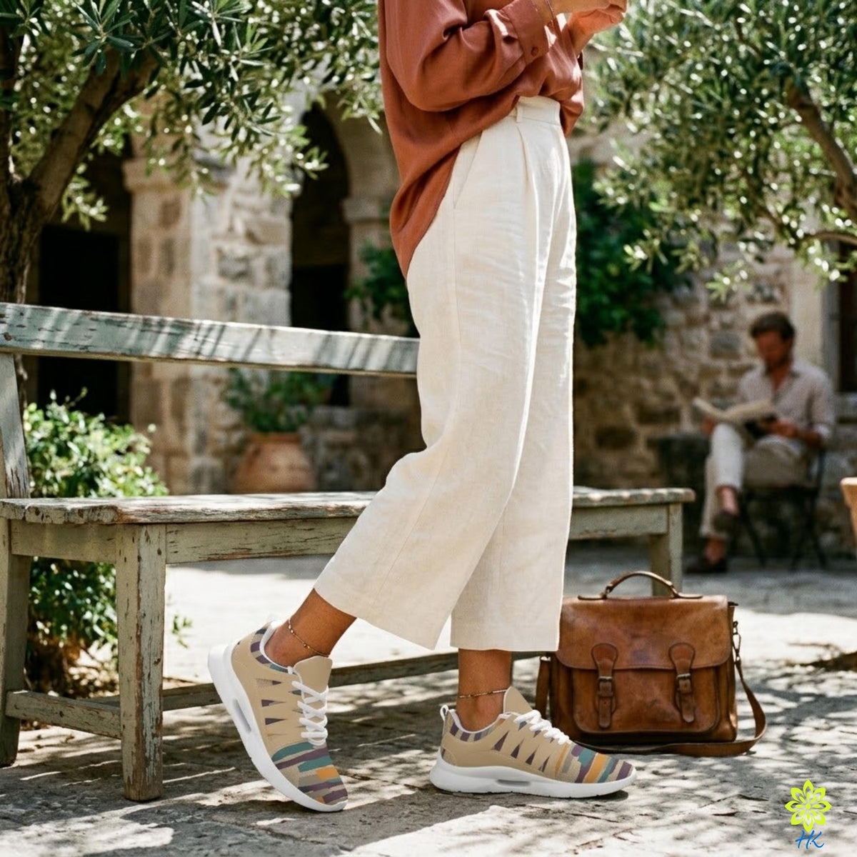 A person wearing a red sweater and white pants stands next to a brown leather bag, with a bench and potted plants in the background.