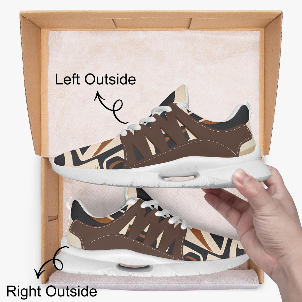 A person's hand is holding a pair of brown and white patterned sneakers, with the left shoe positioned outside the box and the right shoe inside the box.