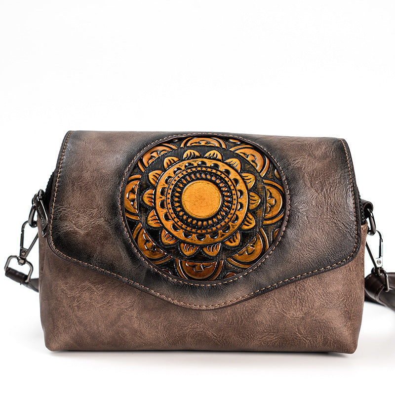 A brown distressed leather crossbody bag featuring a large circular mandala design tooled into the front flap, with a black and gold center and a dark brown outer ring. The bag has a dark brown gradient finish and a black shoulder strap visible on the sides.