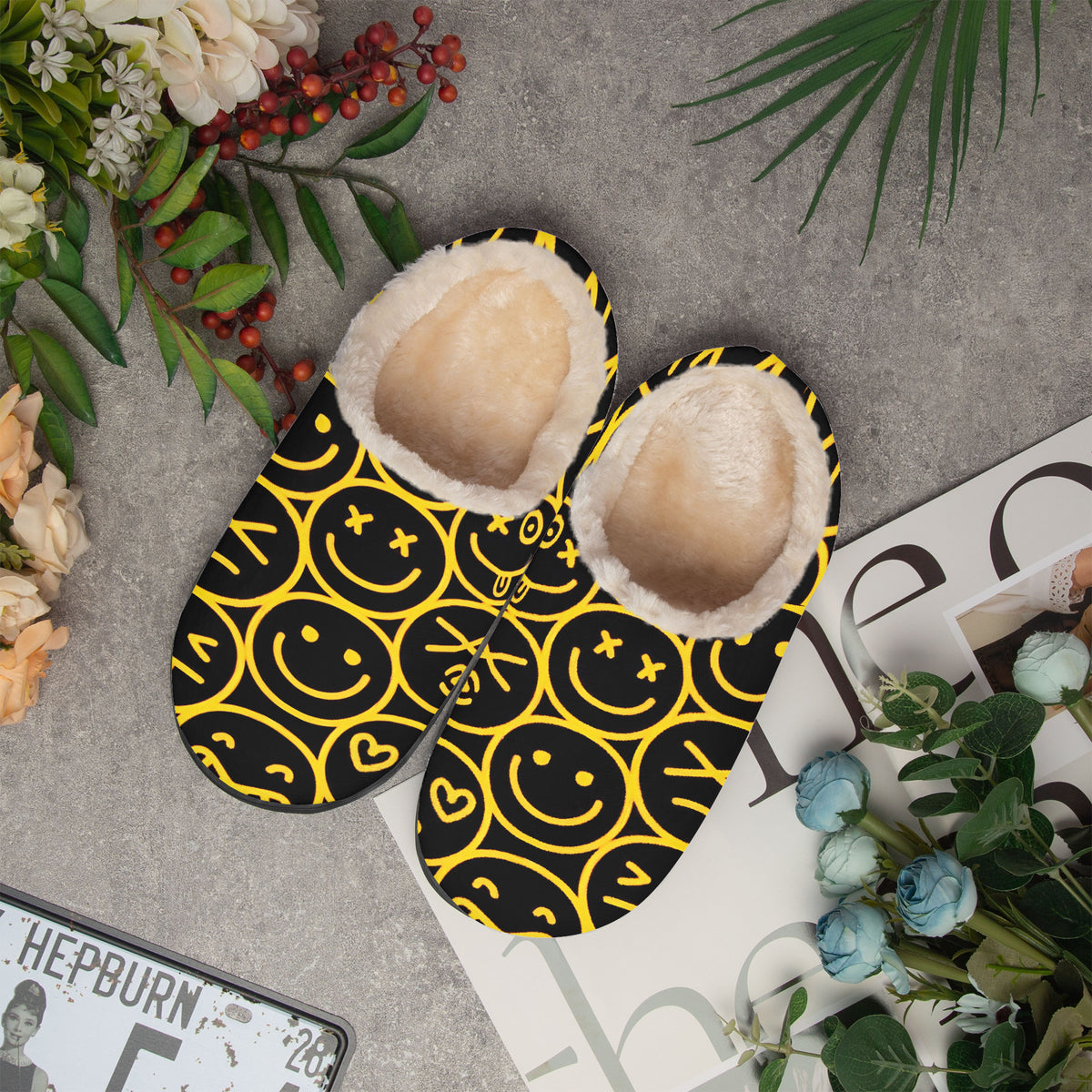 Top view of a pair of smiley face slippers on a grey surface with flowers and a magazine