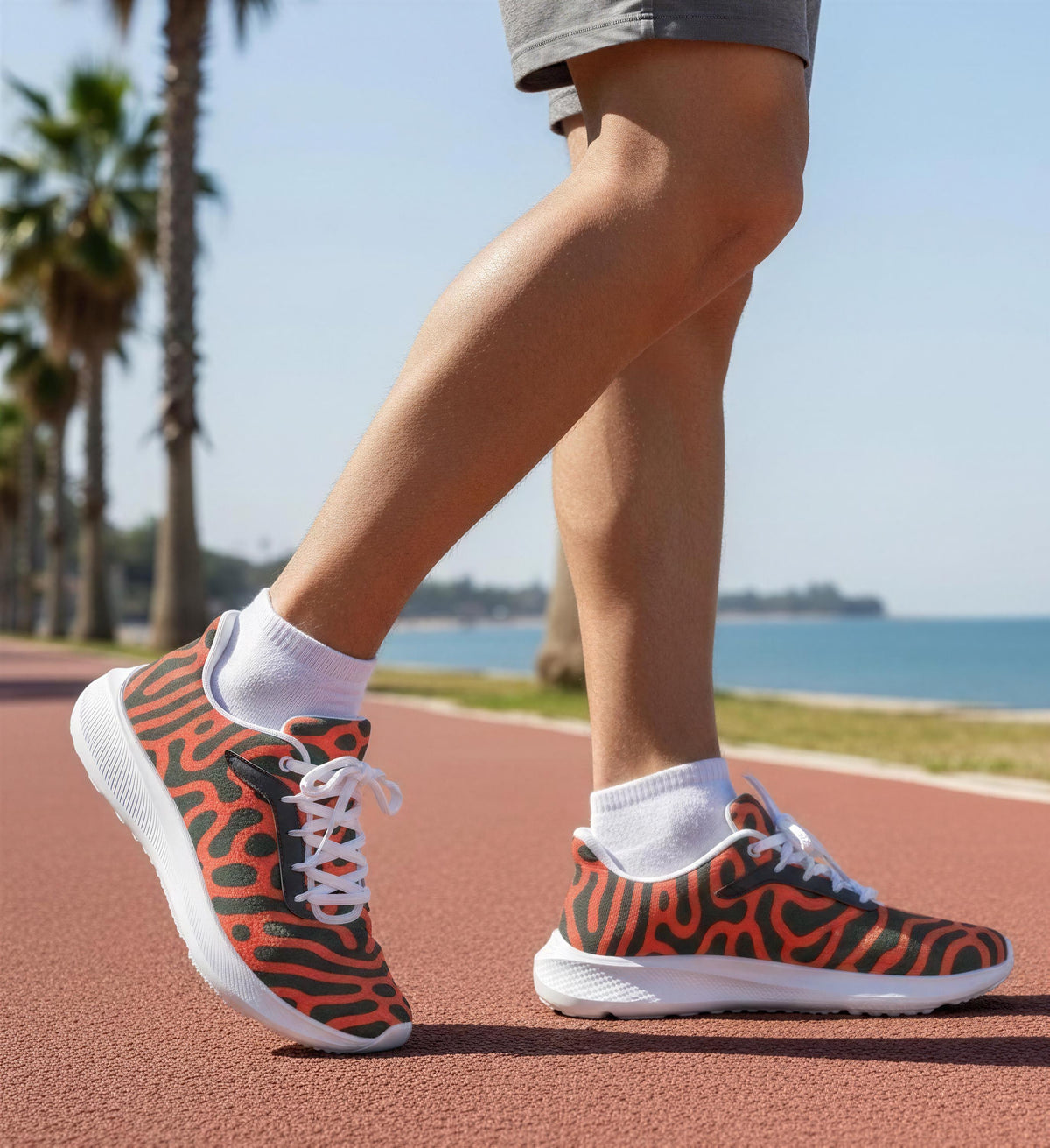 A person wearing a pair of orange and black patterned sneakers is walking on a red track near a beach with palm trees in the background.