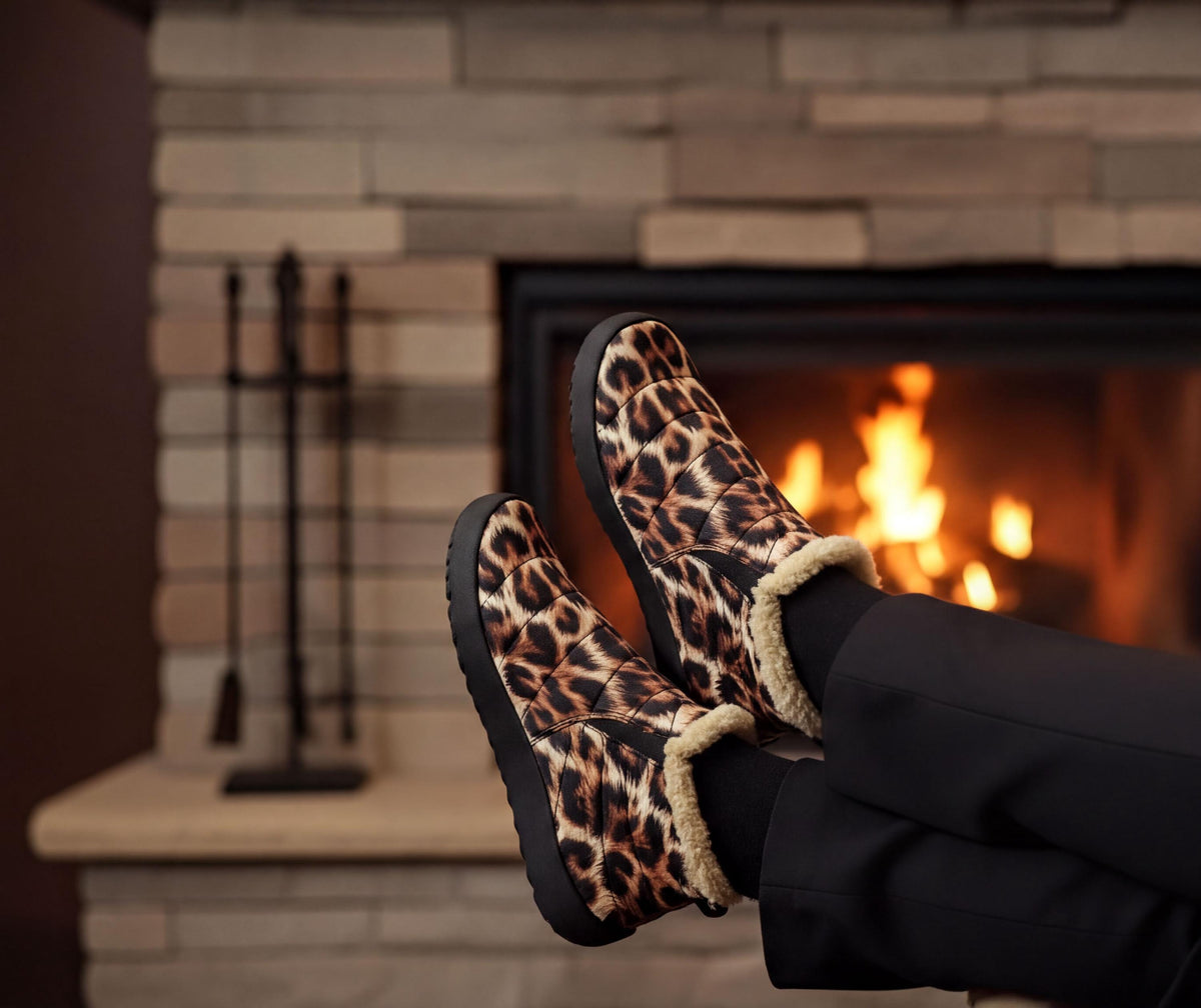 A pair of leopard print boots with fur trim, placed on a person's feet, with a warm fireplace visible in the background.