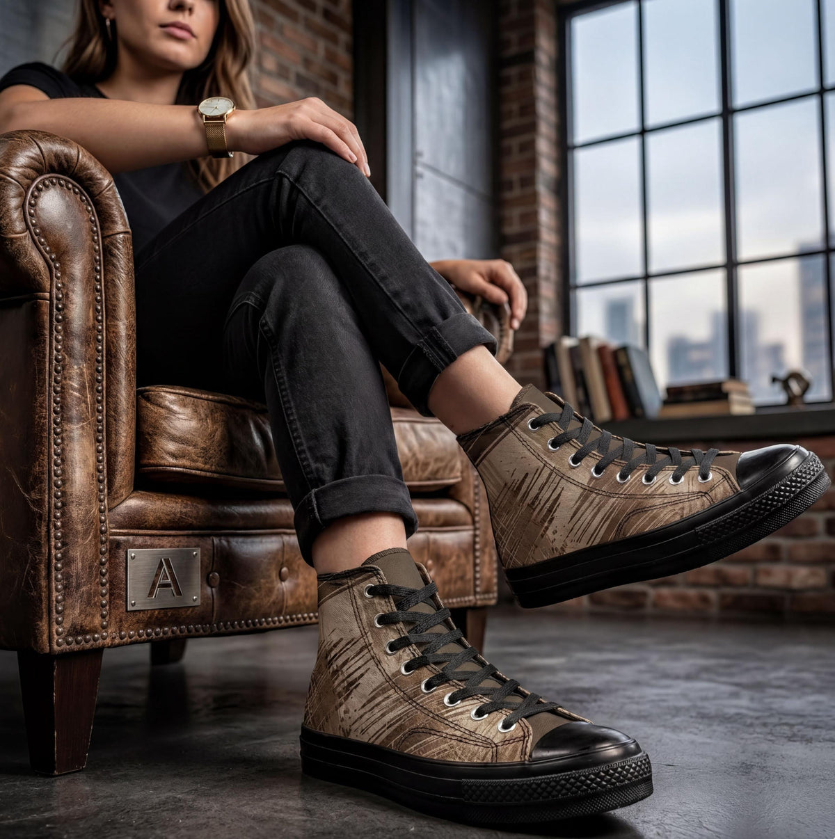 A woman wearing black pants and tan and black sneakers is sitting on a leather chair in a room with a brick wall and a window.