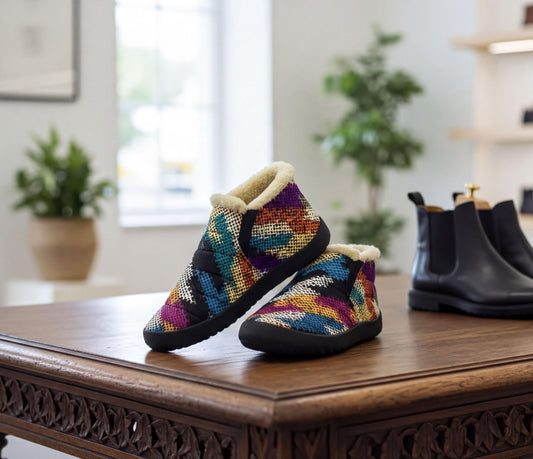 A pair of colorful, patterned slippers with fur lining is displayed on a wooden table, with a pair of black leather boots visible in the background.