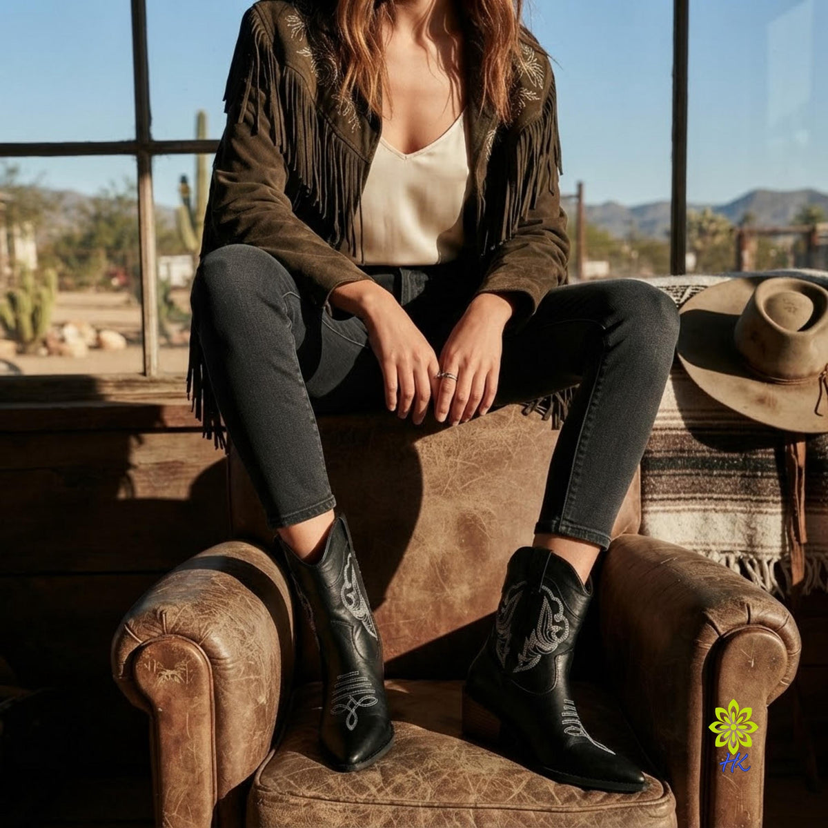 A woman sits on a rustic leather armchair in a western-style room, wearing black fringed suede boots with silver embroidery. She is dressed in a cream top, dark gray jeans, and a brown fringe jacket, with a cowboy hat resting on the chair beside her.