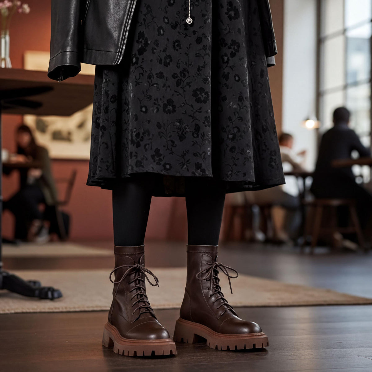 A person wearing a black floral dress and brown lace-up boots stands in a room with other people in the background.