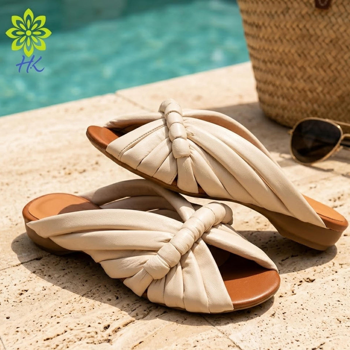 A pair of beige sandals with a twisted, knotted design are placed on a tiled surface near a pool, with a straw hat and sunglasses visible in the background.