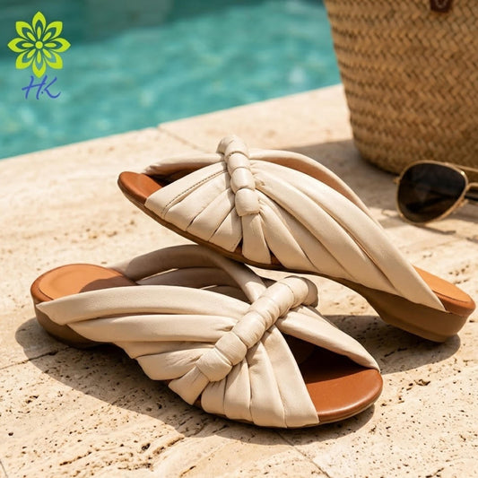 A pair of beige sandals with a twisted, knotted design are placed on a tiled surface near a pool, with a straw hat and sunglasses visible in the background.