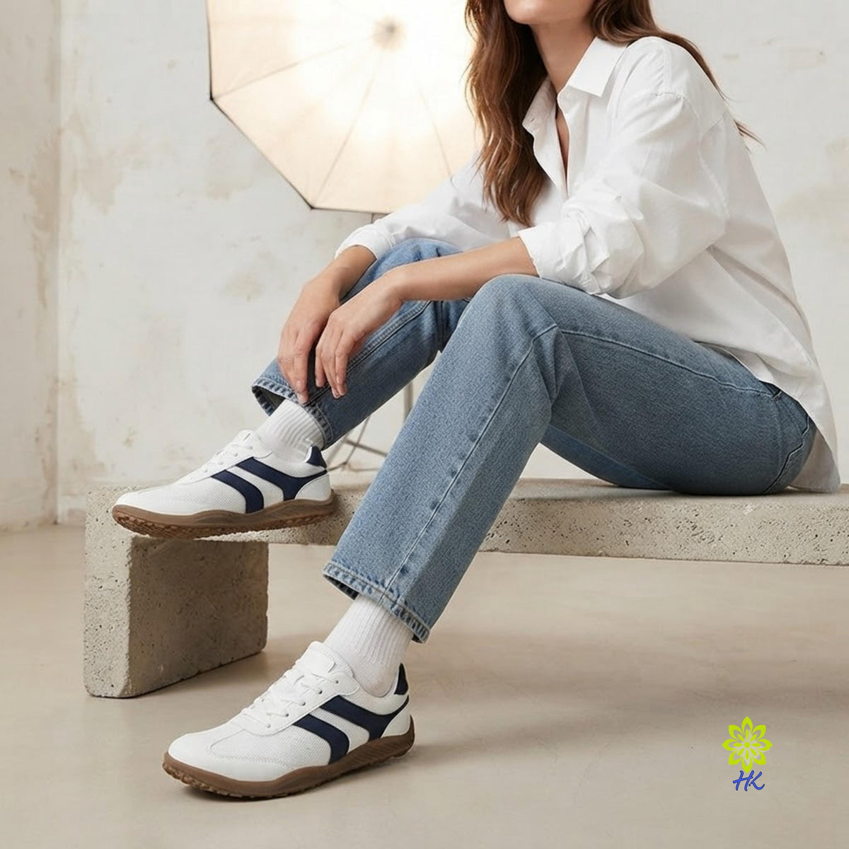 A woman sits on a concrete bench wearing white low-top sneakers with navy blue side stripes, light blue jeans, and white socks in a bright, neutral indoor setting.