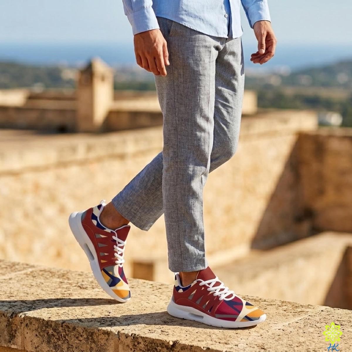 A person wearing gray pants and colorful sneakers stands on a stone ledge, with a scenic view of the ocean and buildings in the background.
