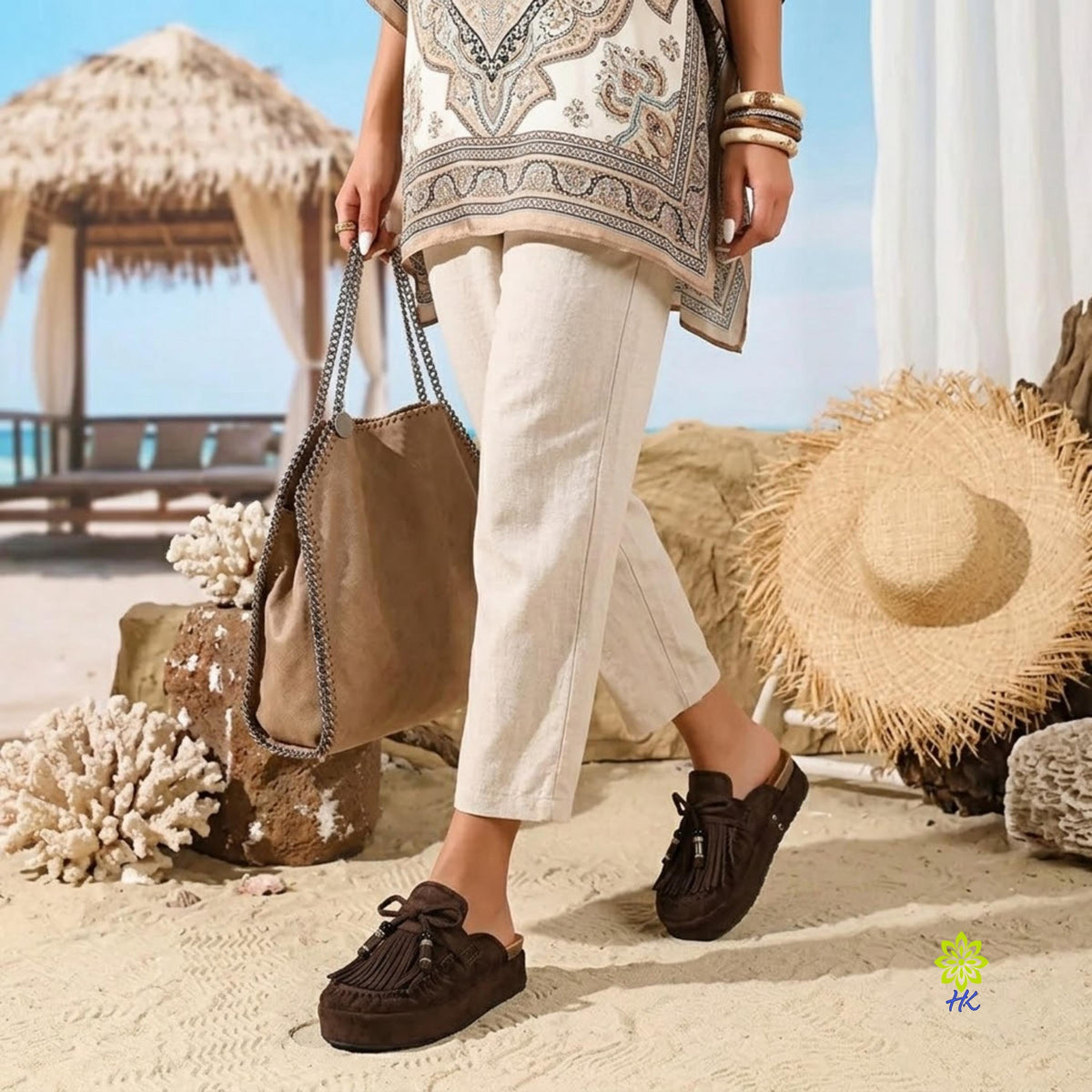 A person walking on a sandy beach wearing brown tassel mule sandals, cream pants, and a patterned beige top, while carrying a taupe tote bag. A straw hat, thatched cabana, and coral are visible in the background.