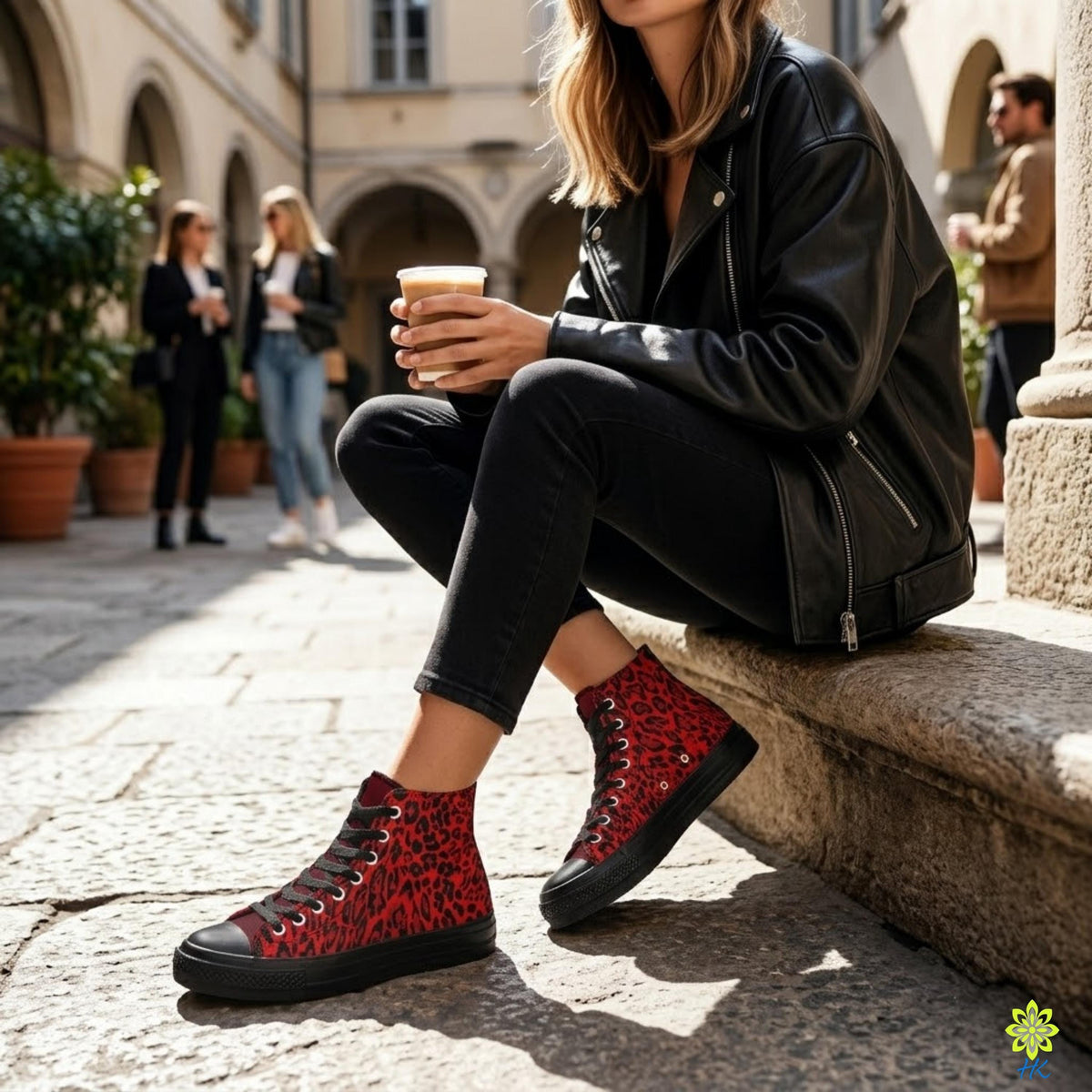 A woman in a black leather jacket and red high-top sneakers is sitting on a stone ledge, holding a coffee cup.