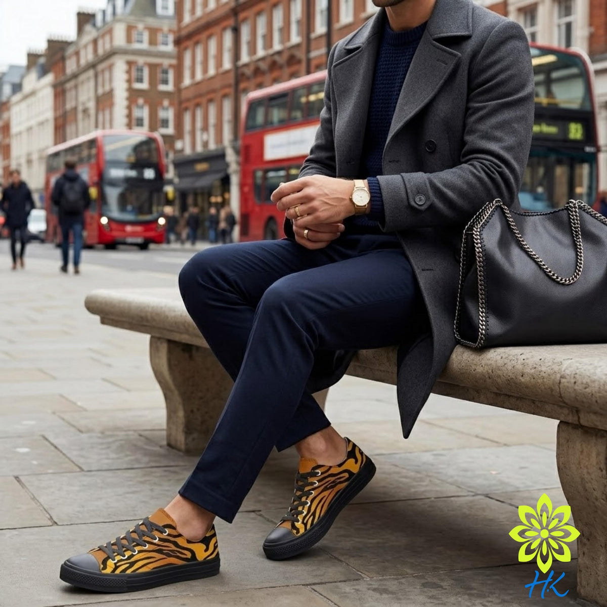 A man in a gray coat and navy blue pants sits on a bench in a city, wearing a watch and holding a black purse.