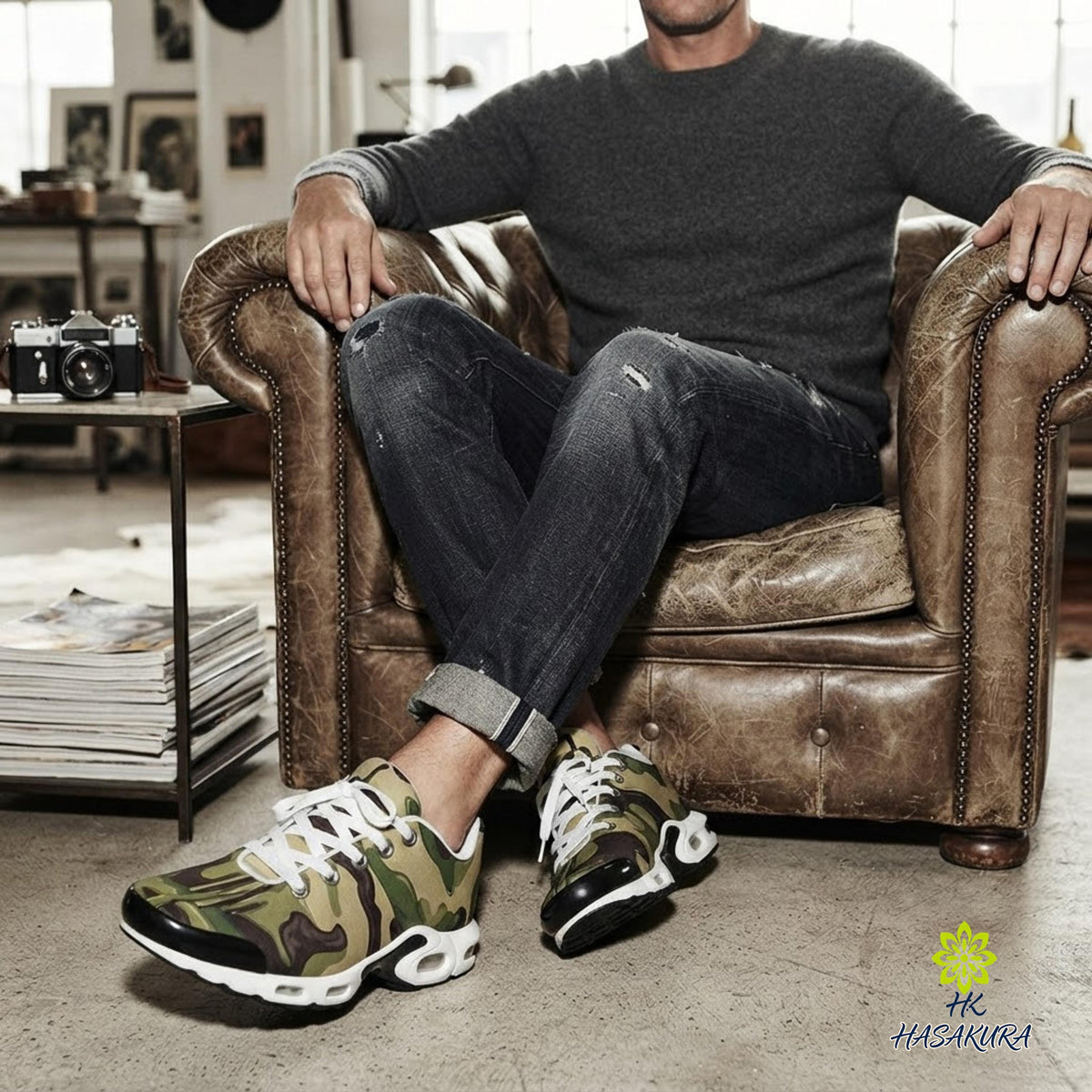 A man wearing a gray sweater and camouflage sneakers is sitting in a leather armchair, with a camera on a table next to him.
