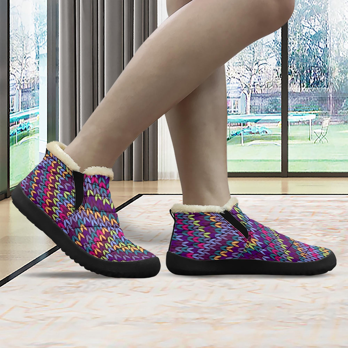 A person's feet wearing colorful, patterned boots with a fur lining, standing in front of a window with a view of a garden outside.