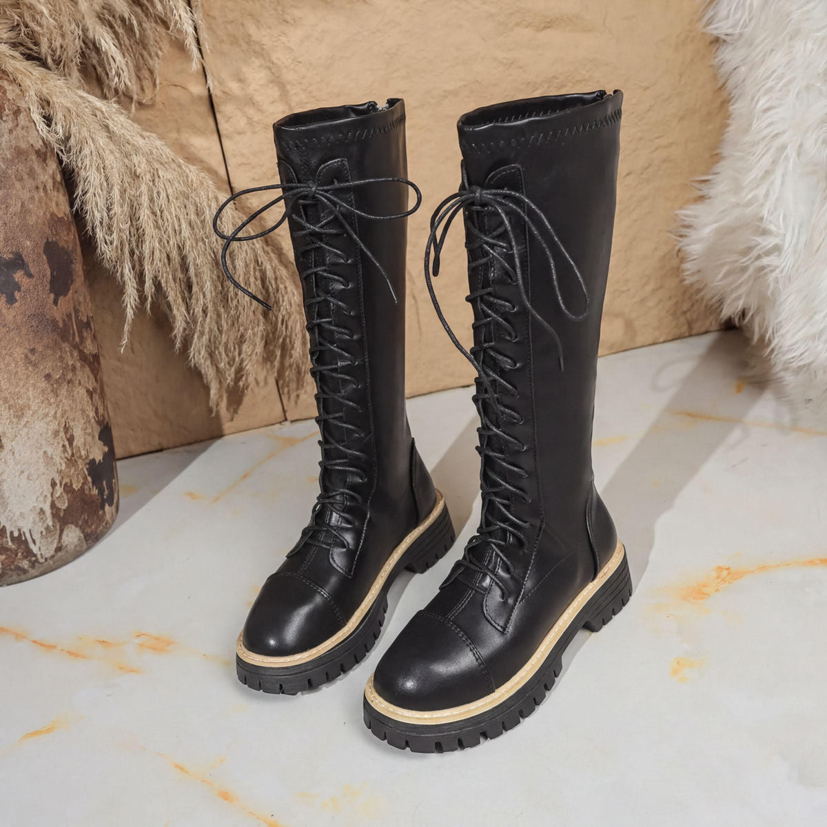 A pair of black leather boots with laces and a chunky sole, placed on a white surface against a beige background.