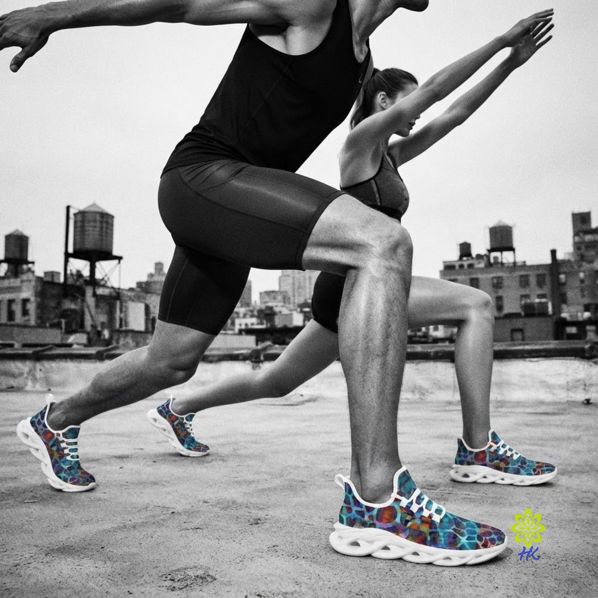 Two dancers in black athletic wear and colorful patterned sneakers perform a low lunge on a rooftop with a city skyline in the background. The photo is in black and white except for the shoes, which show blue, red, and purple patterning.