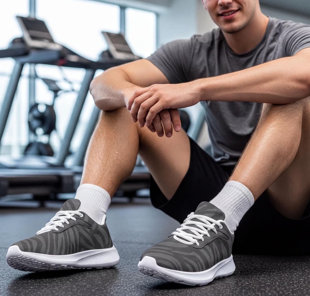 A man is sitting on a bench in a gym, wearing gray and white sneakers, and resting his hands on his knees.