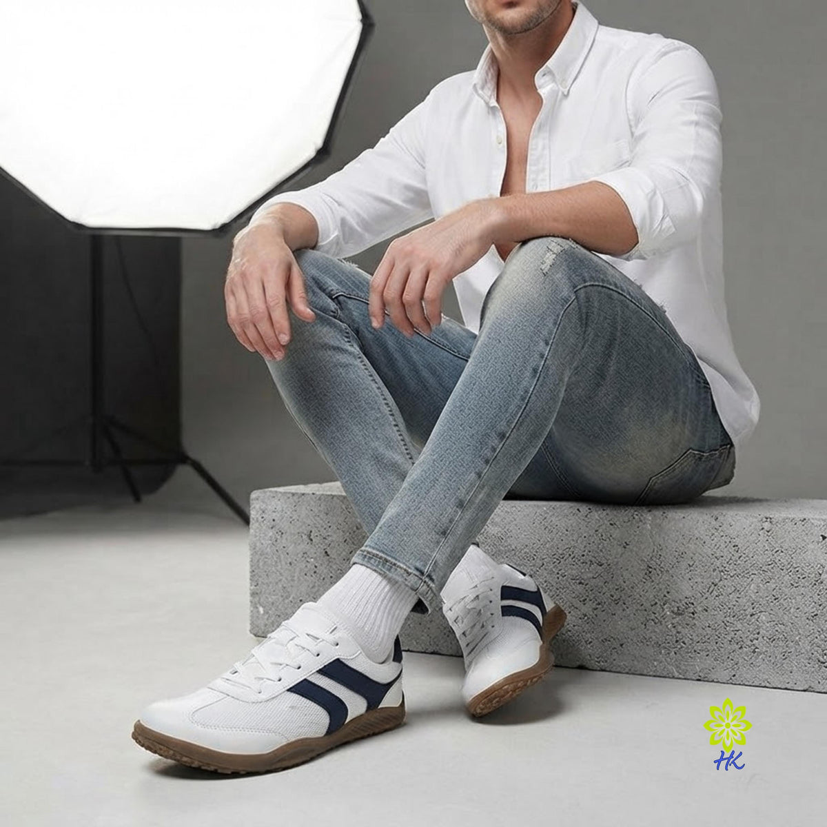 A man wearing a white button-down shirt, light blue jeans, white socks, and white low-top sneakers with navy blue side stripes sits on a concrete block in a studio setting.