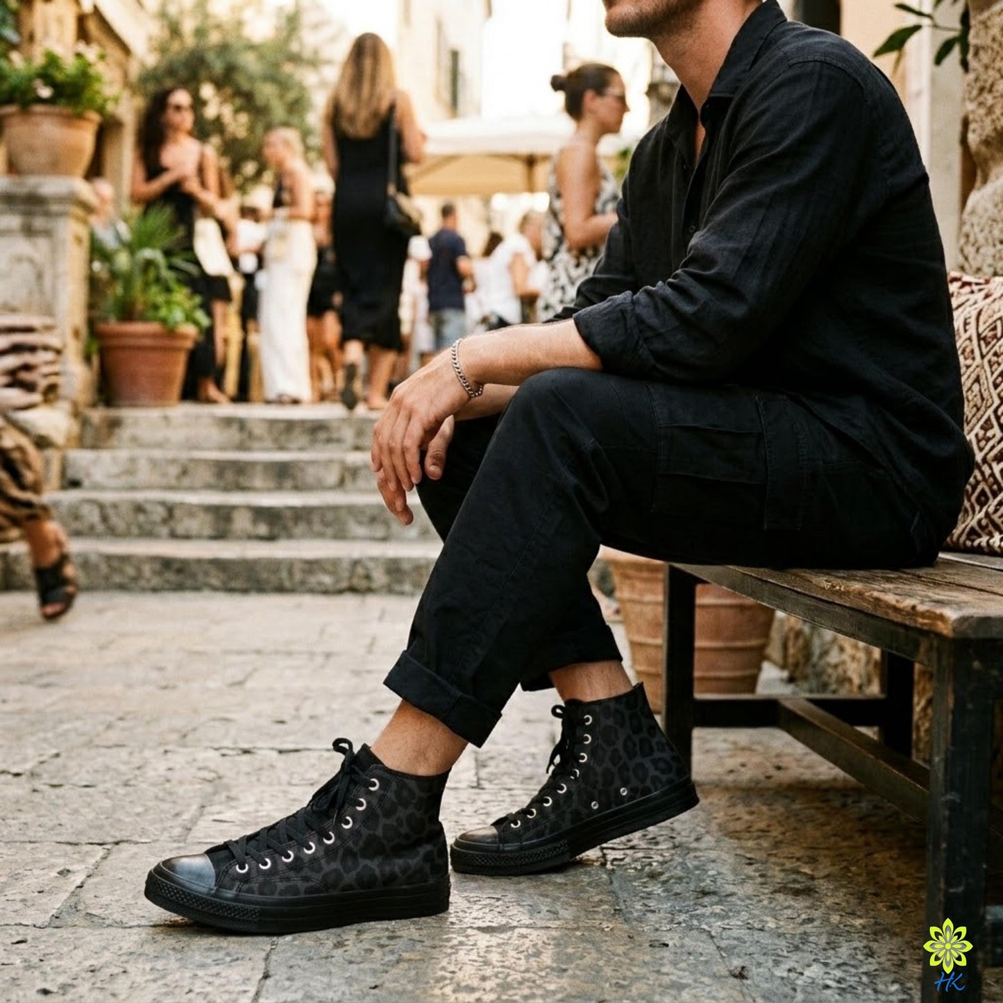 A man wearing black clothing and high-top sneakers is sitting on a bench, with other people visible in the background.