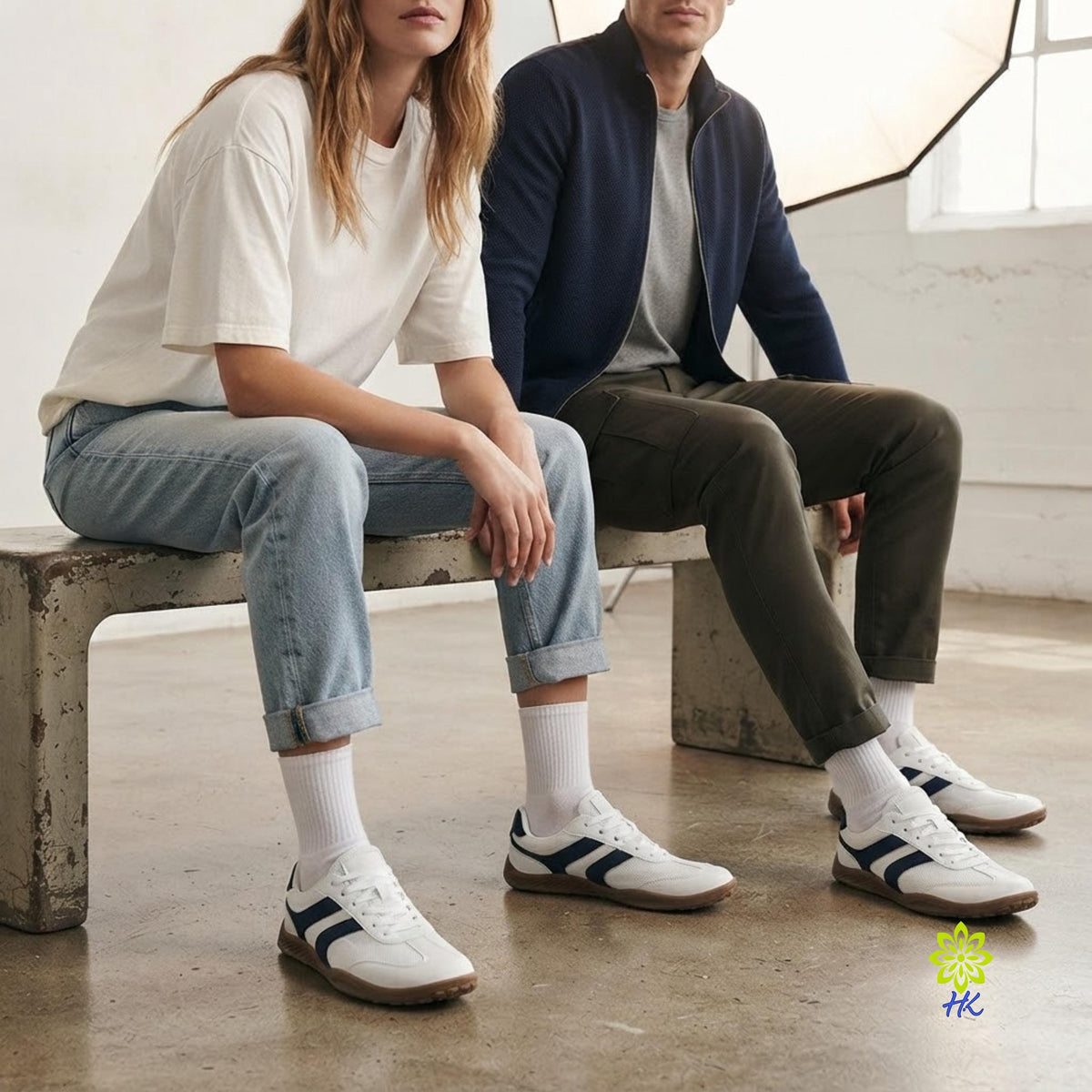 Two models sitting on a concrete bench in a bright studio setting, wearing white low-top sneakers with dark navy stripes, white crew socks, and casual jeans and a jacket.