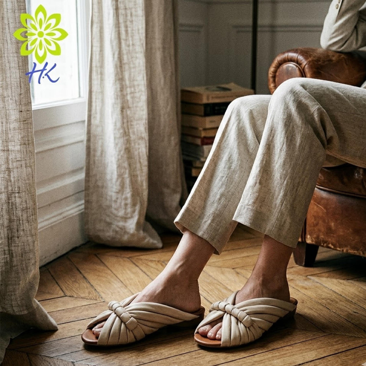 A person is sitting in a chair, wearing beige pants and white sandals, with a window and bookshelf visible in the background.