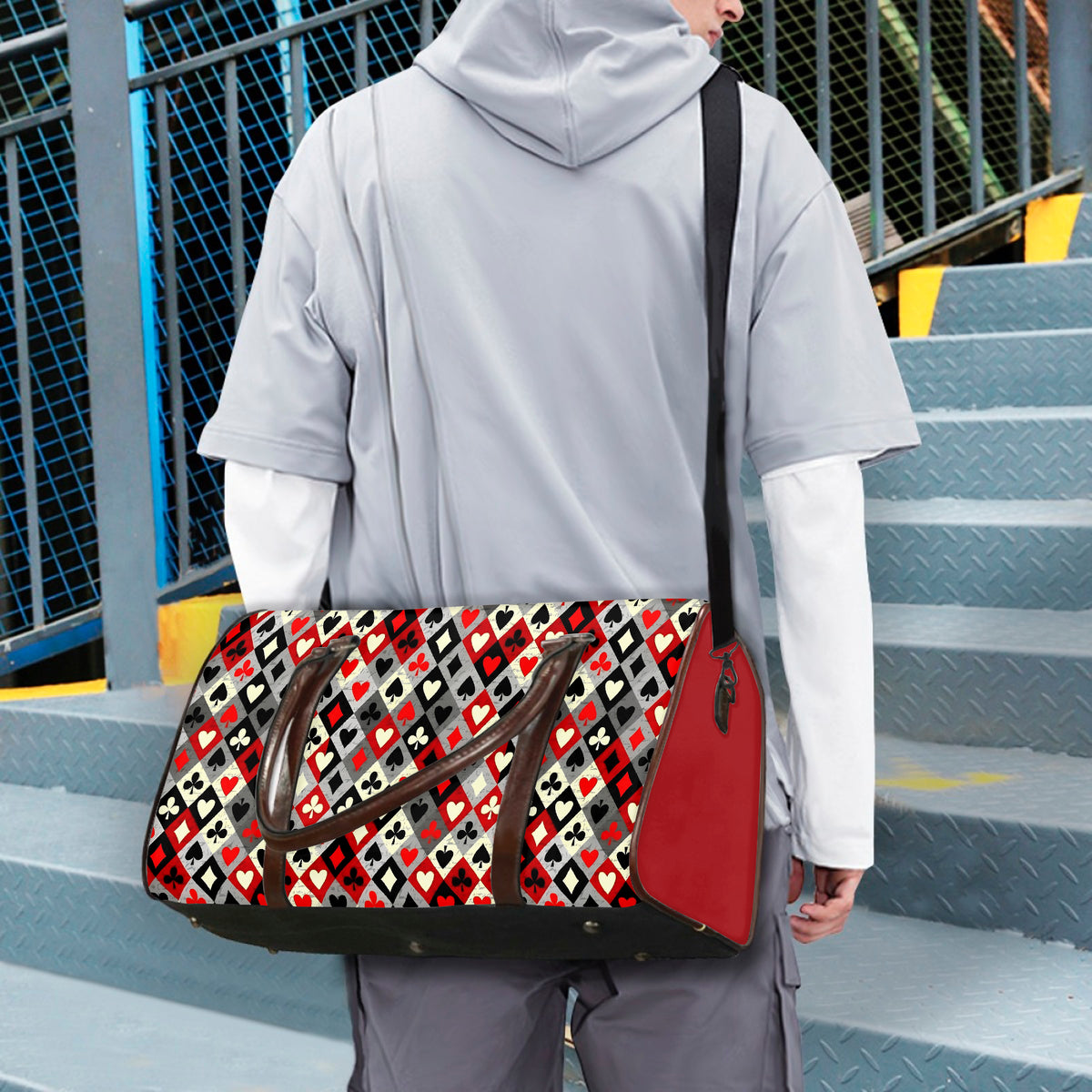 A person wearing a light gray hoodie and white long sleeves carries a large black and red sports bag with a geometric heart pattern and brown handles, standing on metal stairs.