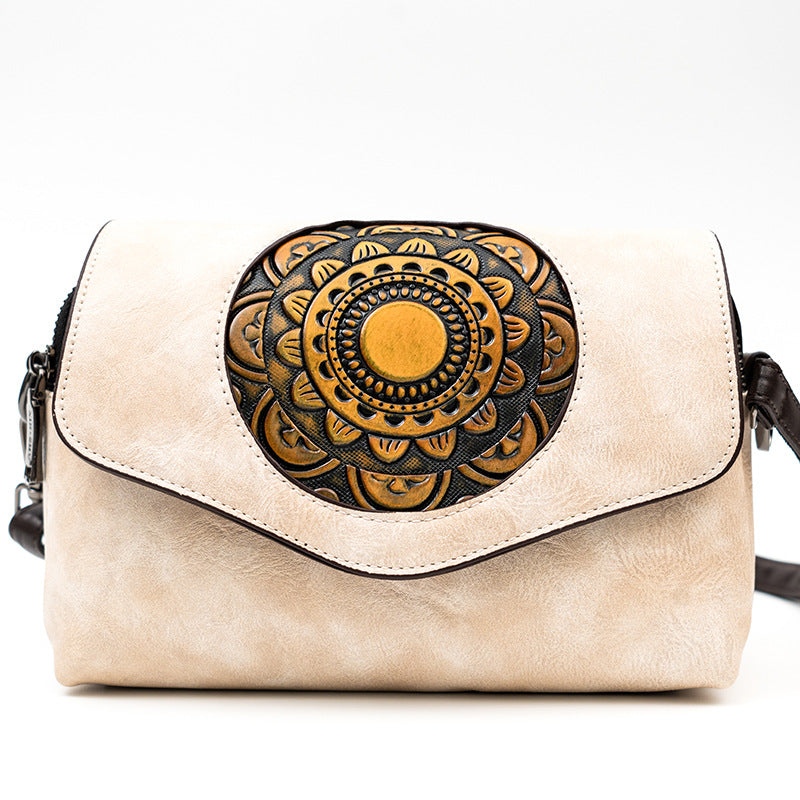 A beige faux-leather crossbody bag with a large circular black and gold hand-tooled-style mandala design on the front flap, shown against a plain white background.