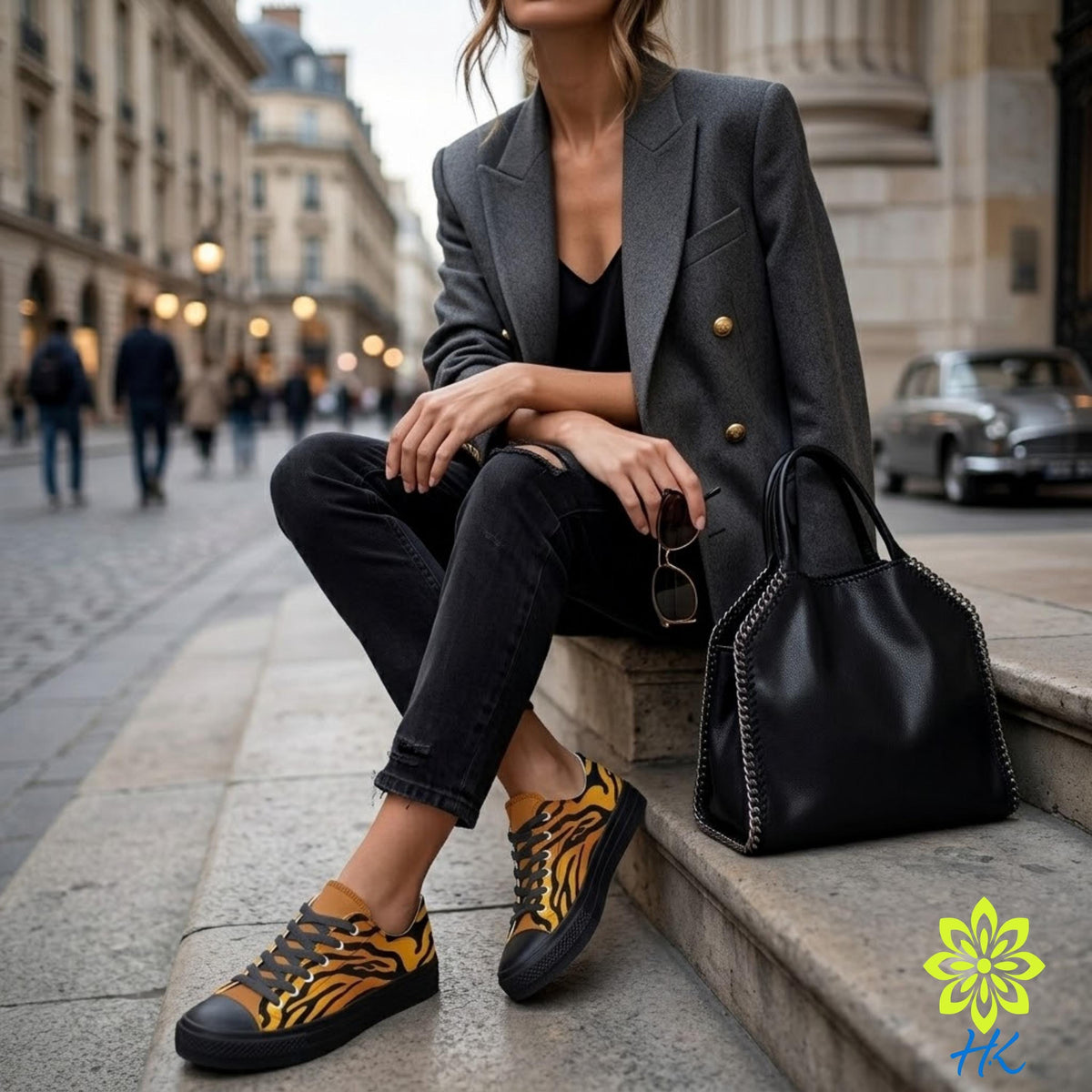 A woman in a gray blazer, black pants, and yellow and black sneakers sits on a stone step, holding a black handbag and wearing sunglasses.