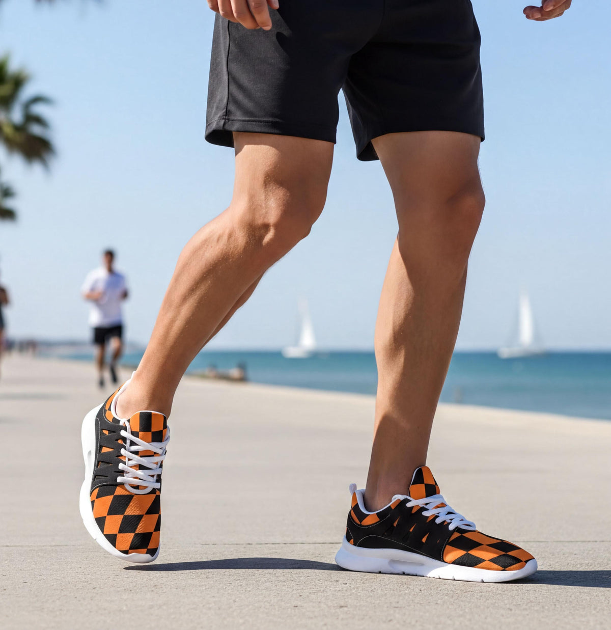 A person wearing black shorts and orange and black checkered sneakers is walking on a beach with sailboats in the background.