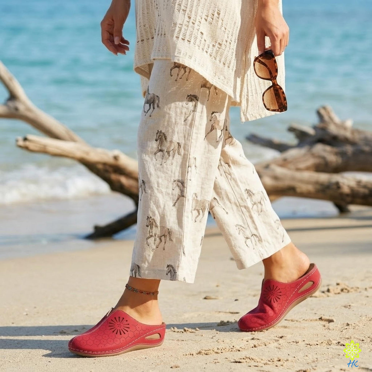 A person wearing a white top and beige pants with a pattern of horses walking on a beach, carrying a pair of sunglasses.