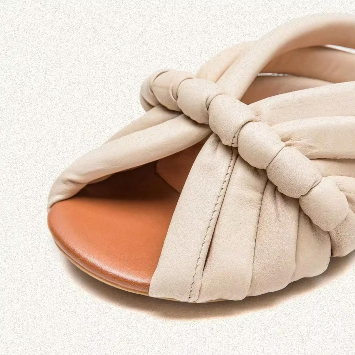 A close-up image of a beige leather sandal with a twisted, knotted design.