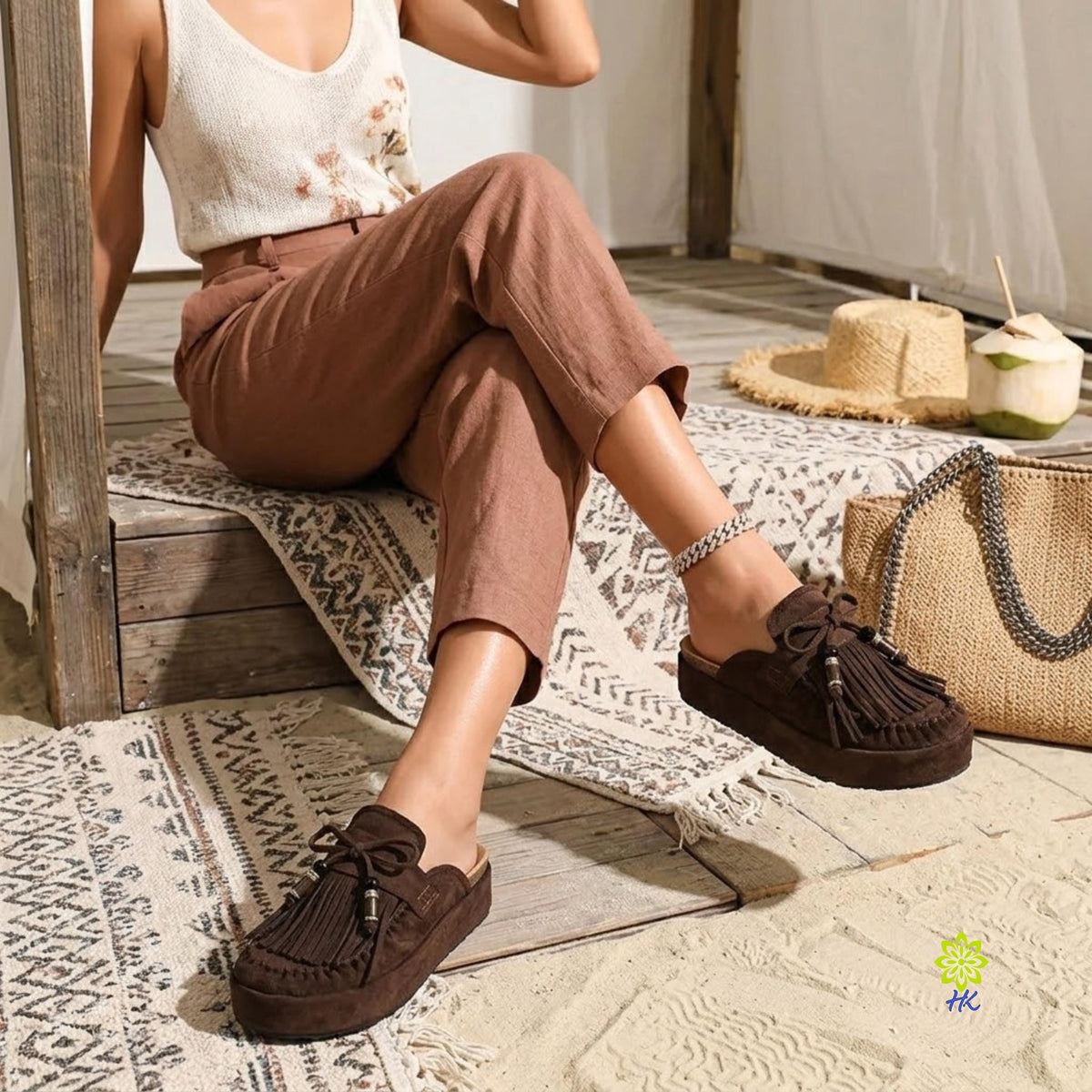 A woman seated on a wooden bench wearing brown suede tassel mule loafers, brown cropped pants, and a white sleeveless top in a rustic indoor setting with a woven bag, hat, and patterned rug nearby.