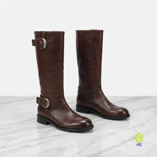 A pair of dark brown leather knee-high boots with silver buckle straps, displayed on a light gray marble surface against a white background.