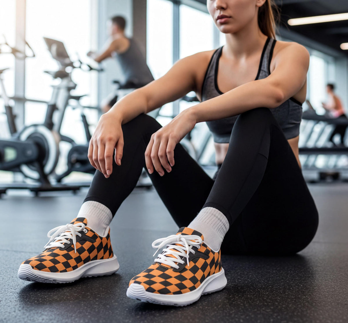 A woman in athletic clothing sits on the floor of a gym, wearing orange and black checkered sneakers.