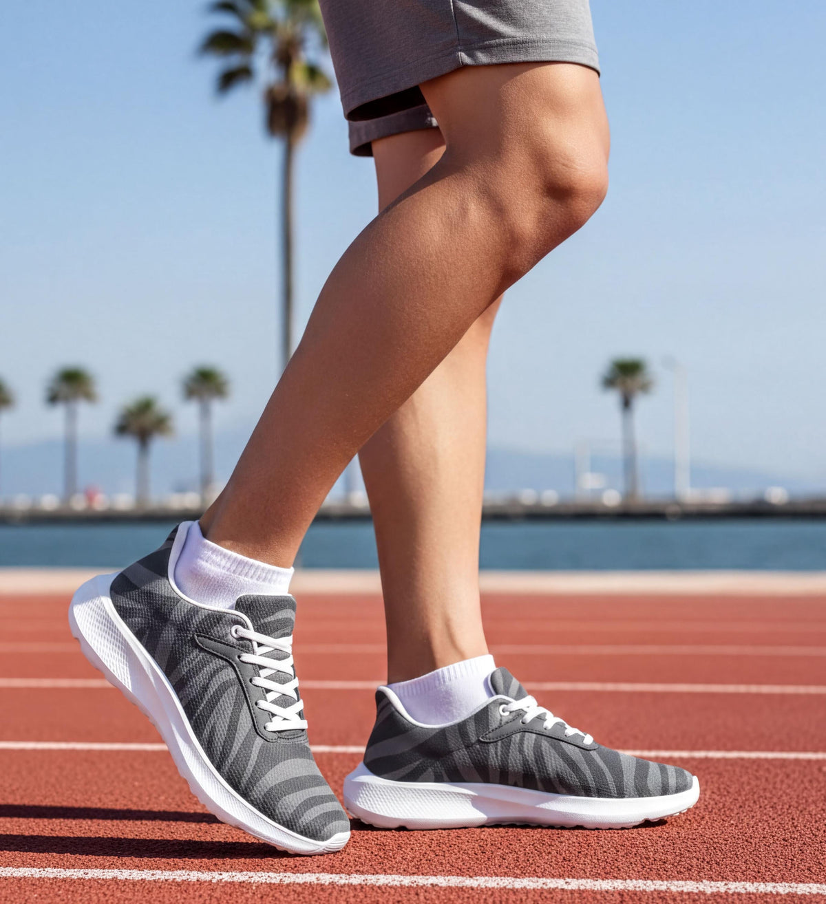 A person wearing gray and white sneakers stands on a red running track, with palm trees and a body of water visible in the background.
