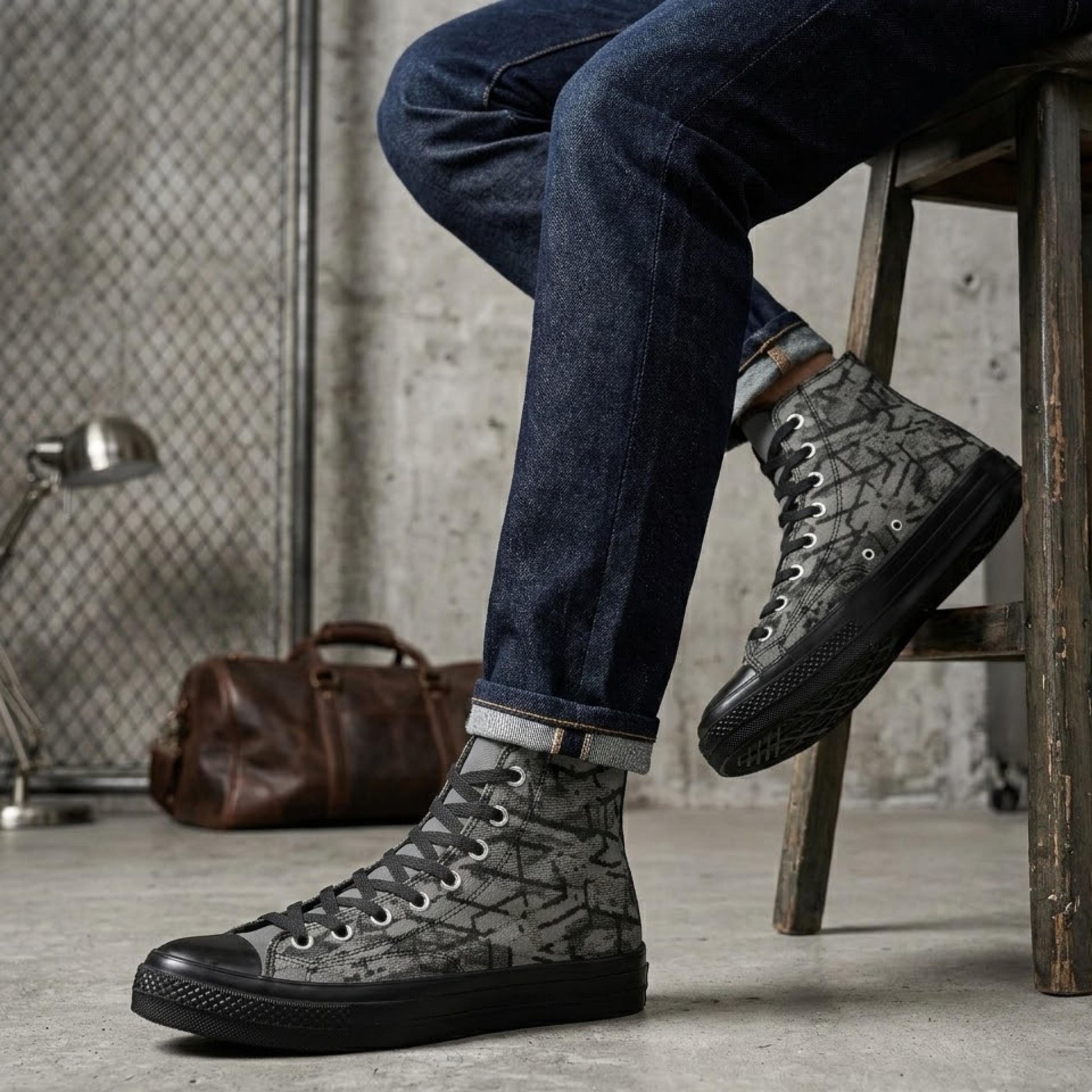 A person is sitting on a wooden stool, wearing black and gray patterned sneakers and blue jeans.