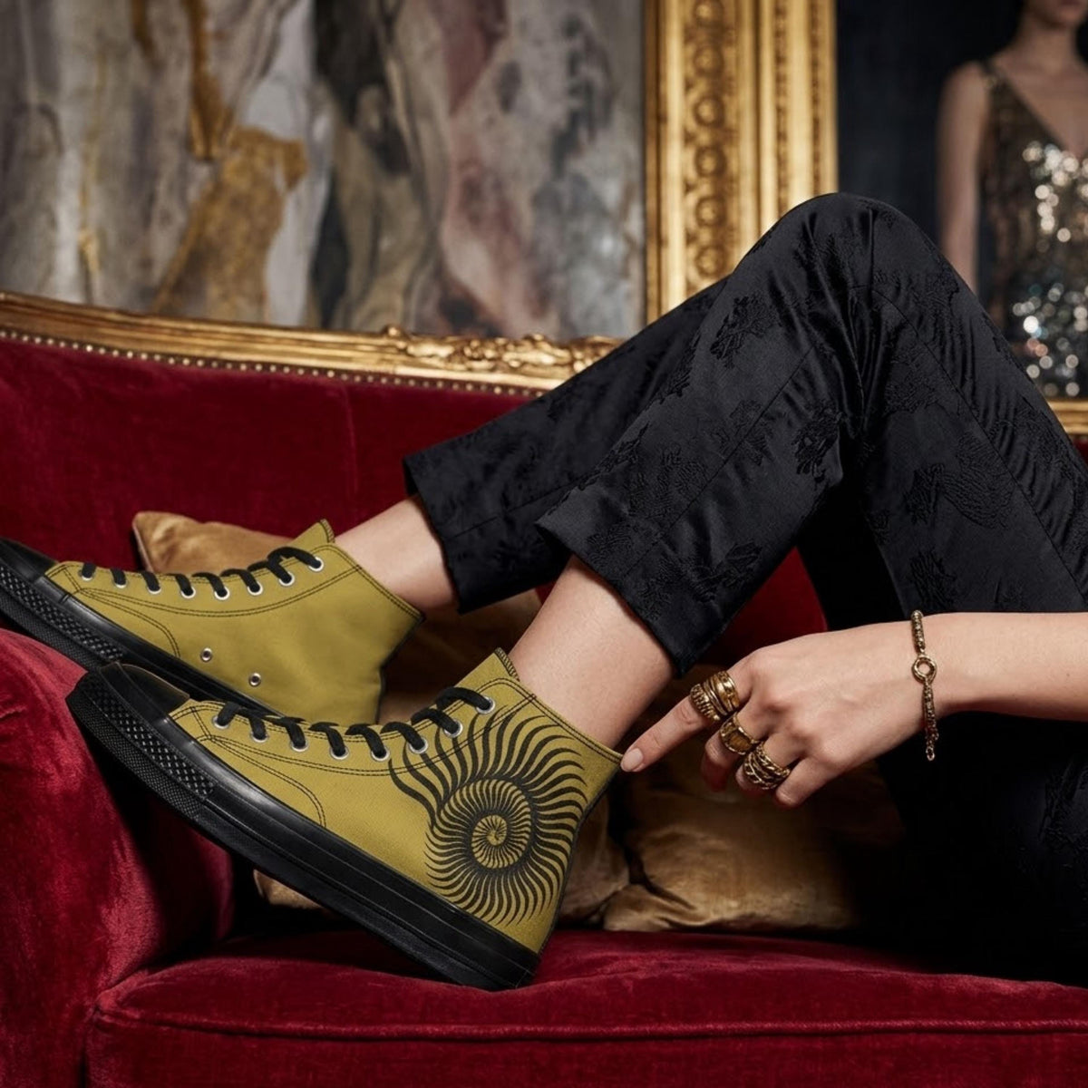 A person is sitting on a red velvet couch, wearing yellow high-top sneakers with a black and gold design, and accessorized with rings and bracelets.