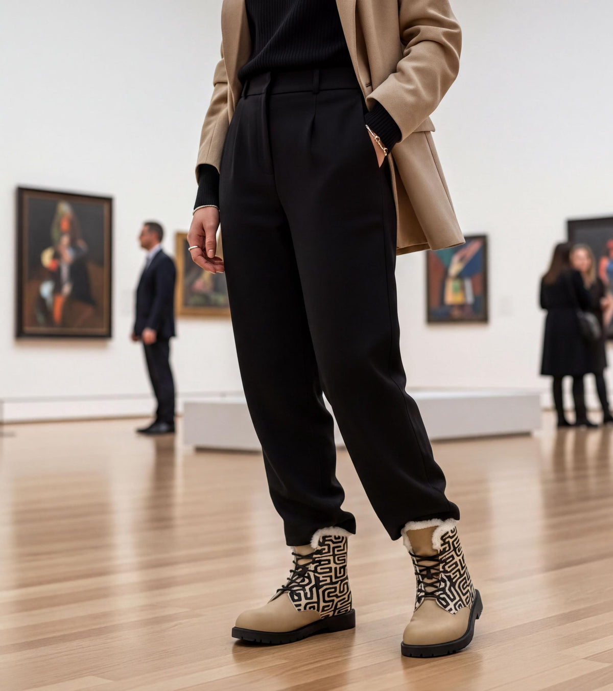 A person wearing a beige jacket, black pants, and patterned boots stands in an art gallery, surrounded by other people and paintings on the walls.