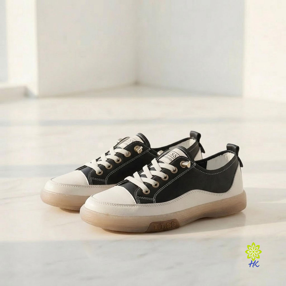 A pair of low-top sneakers in black and off-white canvas, featuring white laces and a translucent gum sole, displayed on a light marble floor in a bright room.