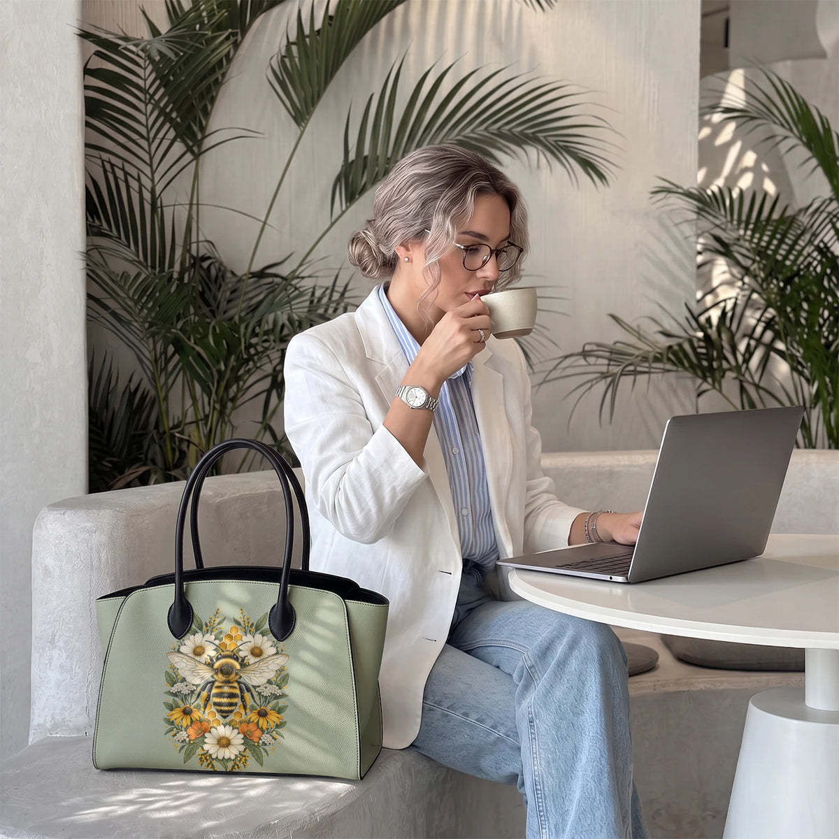 Donna seduta a un tavolo con una borsa tote bee verde salvia accanto a un laptop e a un caffè