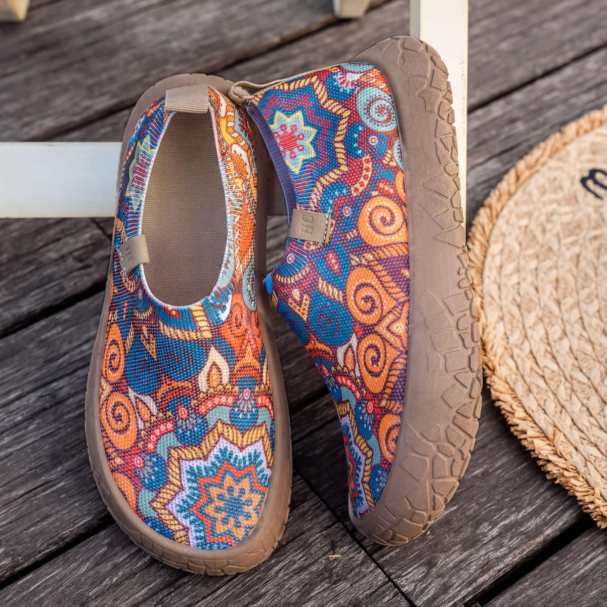 A pair of colorful, patterned shoes with a floral design, placed on a wooden surface next to a straw mat.