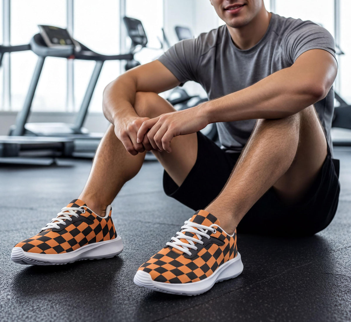 A man is sitting on the floor in a gym, wearing orange and black checkered sneakers.