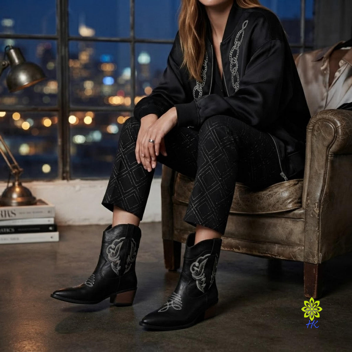 A model sits in a leather armchair wearing a matching black western-style outfit with embroidered details and black pointed-toe boots. The setting is a dimly lit room with a window showing a blurred city skyline at night.