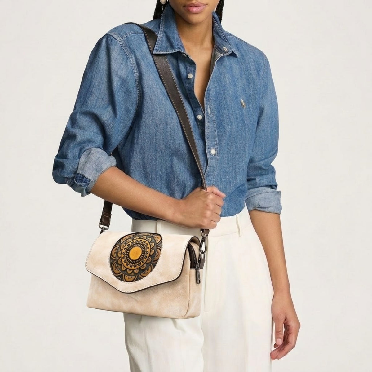 A model wearing a blue denim shirt and white pants carries a cream-colored crossbody bag with a brown strap and a large circular black and gold mandala pattern on the front flap.