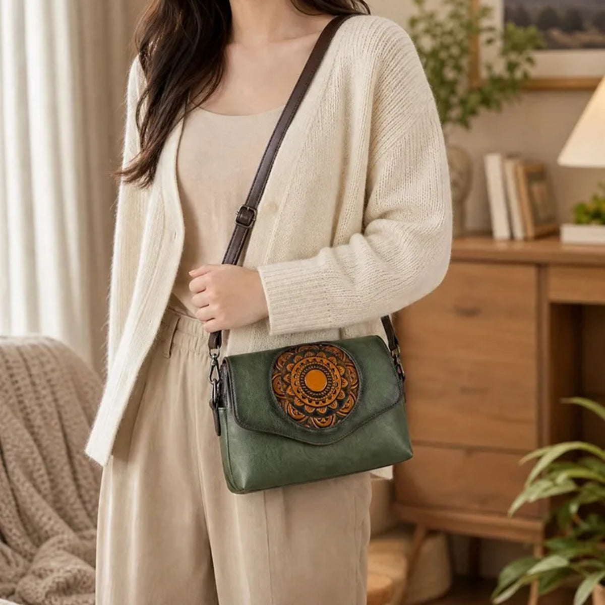 A woman wearing a cream-colored cardigan and matching pants carries a green crossbody bag with a brown strap and a circular orange floral design on the front, set in a warm, cozy room with wooden furniture and plants.