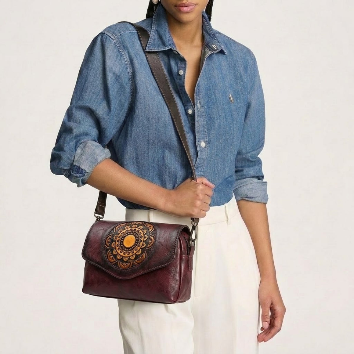 A woman wearing a denim shirt and white pants carries a dark burgundy leather crossbody bag with a brown shoulder strap and a large tan floral design on the front flap.