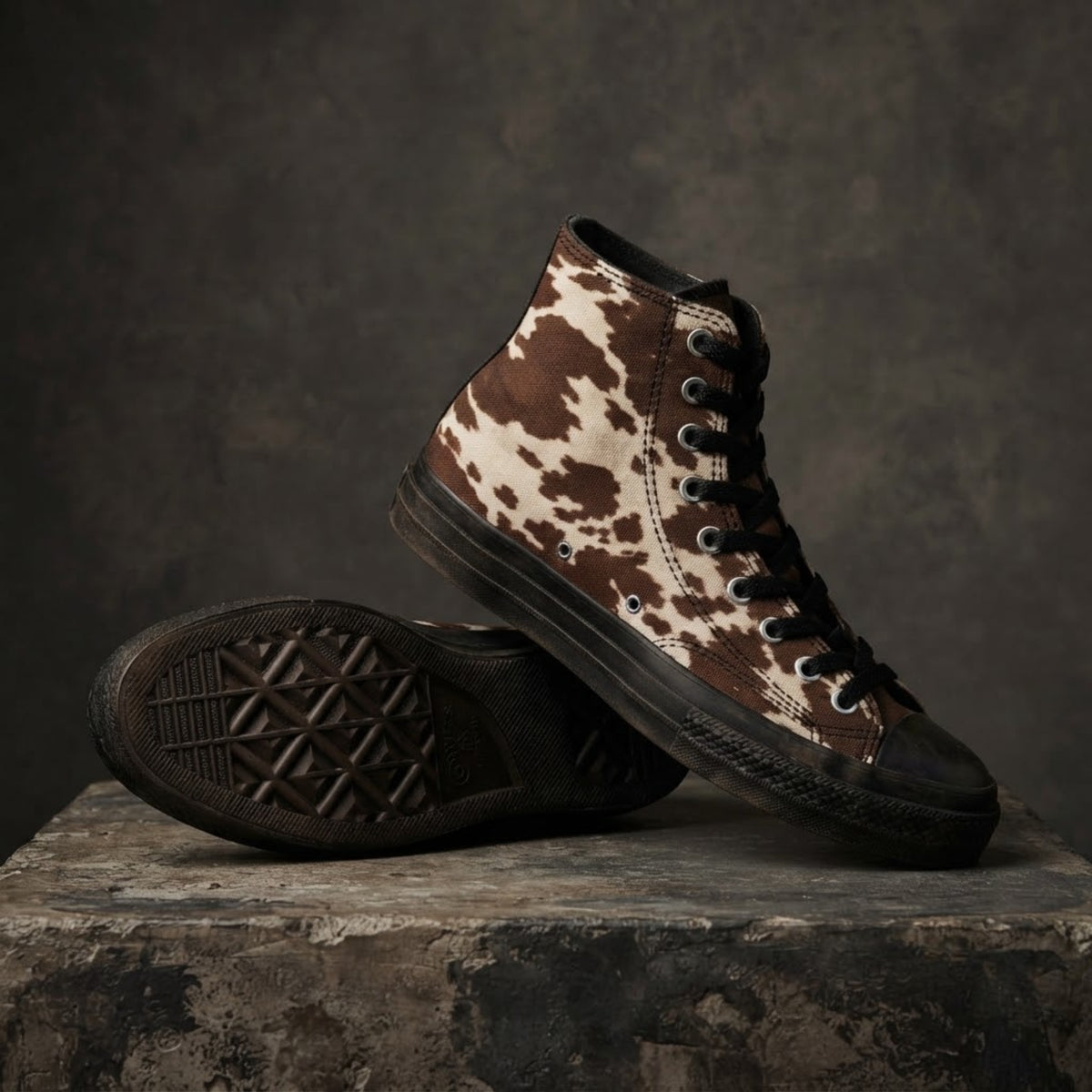 Sneakers alte a stampa mucca con macchie marroni irregolari su tela color crema, mostrate su un piedistallo di pietra grigia