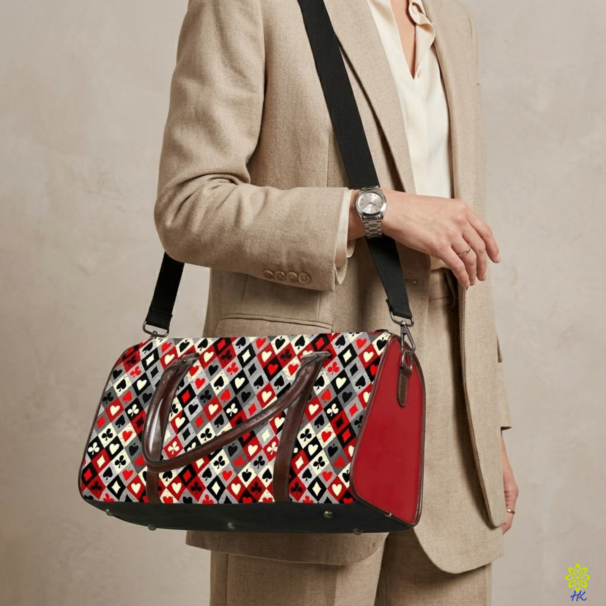 A person in a beige blazer and white blouse carries a patterned duffel bag with a black shoulder strap. The bag features a red, black, white, and gray geometric heart pattern, a solid red side pocket, and dark brown leather handles.