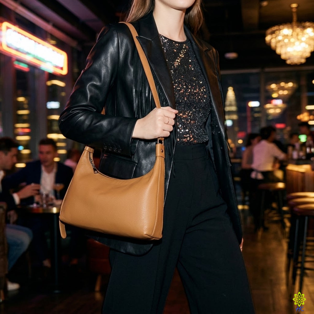 A woman in a black leather jacket and black pants carries a tan leather shoulder bag in a dimly lit, upscale bar or restaurant setting.